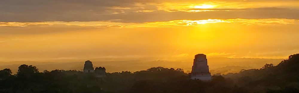 Tikal's Temples IV, I, and II rising out of the jungle silhouetted by a brilliant orange sunrise.